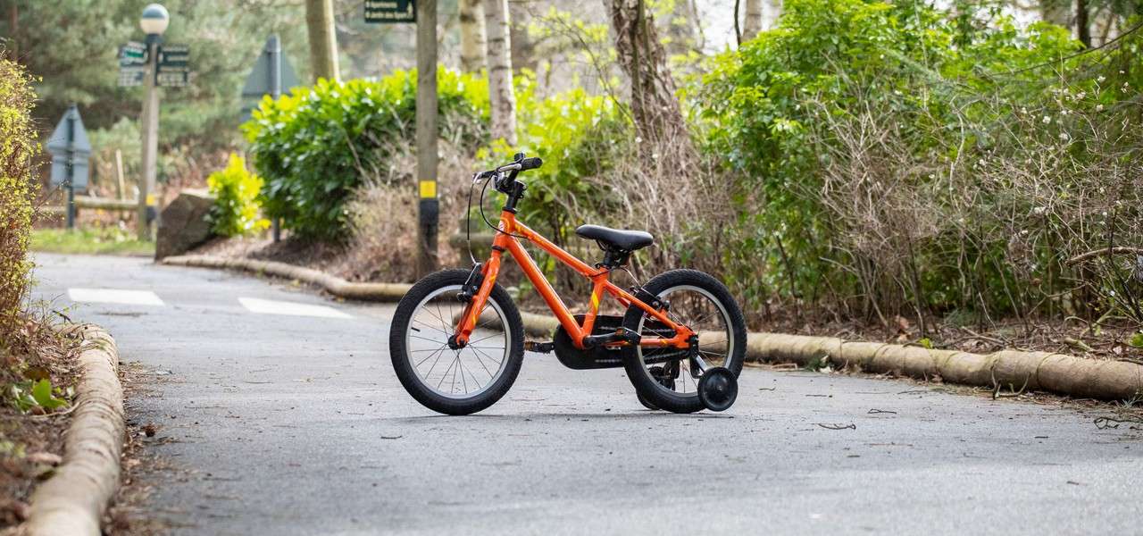 Child’s orange bicycle with training wheels stands idle, slightly angled, on a paved path; surrounded by bushes and trees, with signposts and crosswalk markings in a quiet park.