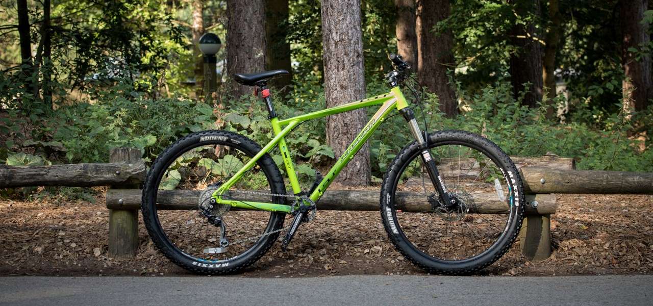 Lime-green hardtail mountain bike leans against a wooden fence. Surrounding trees and shrubs create a forest setting beside a paved path. Text: ARDENT, MAXXIS, MAXXIS.