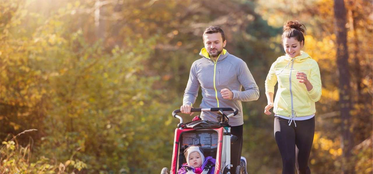 Couple jogs as a man pushes a stroller carrying a bundled toddler; woman runs beside. They move along a sunlit path through autumn trees in a park.