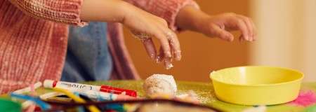 Child’s hands knead a small dough ball, sprinkling powder. Around them, a yellow bowl, markers, and craft materials rest on a green table in a warm indoor setting.