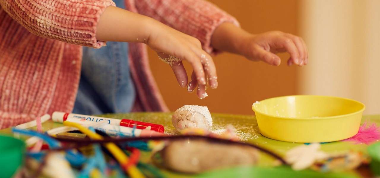 Small hands sprinkle white grains onto a dough ball, on a cluttered craft table with a yellow bowl, markers, pipe cleaners, and feathers.
