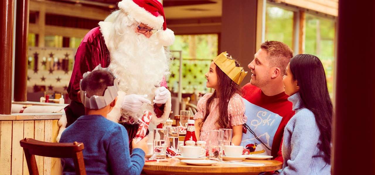 Santa Claus greets children at a dining table, handing a gift. A family in paper crowns watches and smiles amid cups, plates, and festive decorations inside a warmly lit restaurant.