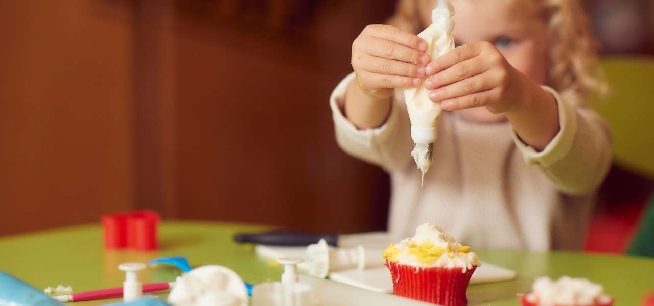 Child squeezes a piping bag, applying white frosting onto a cupcake in a red liner, on a messy green table with decorating tools and ingredients in a warm indoor setting.