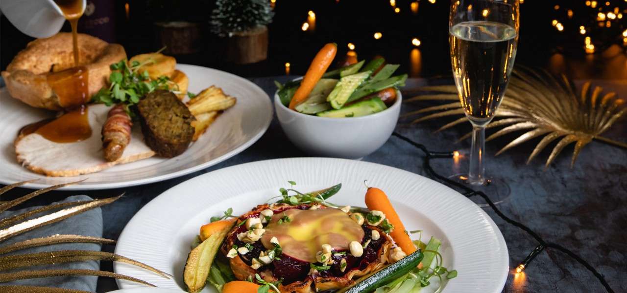 Plated holiday meal sits arranged; gravy is being poured onto a roast with Yorkshire pudding; vegetables and tart accompany; champagne glass and festive lights decorate a dinner table.