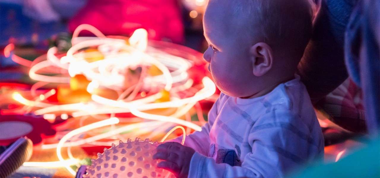 Baby holding a textured spiky ball, gazing at glowing light strands in a dim room, seated on a colorful playmat beside an adult.