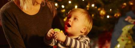 Child holds small toy and looks up, mouth open, toward an adult beside them. Warm lights twinkle on a decorated Christmas tree in the background, suggesting a festive indoor setting.