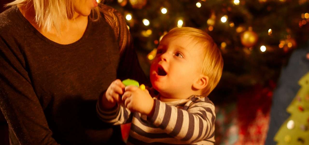 Toddler holds a colorful toy and looks up, mouth open. Seated on an adult’s lap, surrounded by warm lights and ornaments from a Christmas tree in a cozy room.