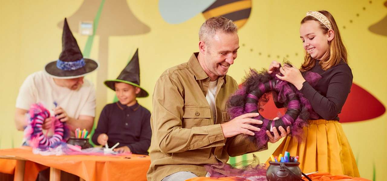 Father and daughter craft a purple-and-black wreath, smiling and holding it together. In a decorated classroom, other participants wear witch hats at orange-covered tables with art supplies.