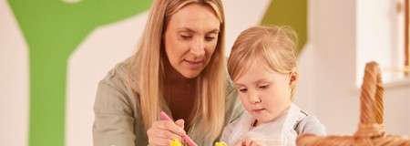 Adult guides child painting with a brush, focusing on a small craft project at a table in a bright playroom, with soft lighting and colorful geometric shapes on the wall.