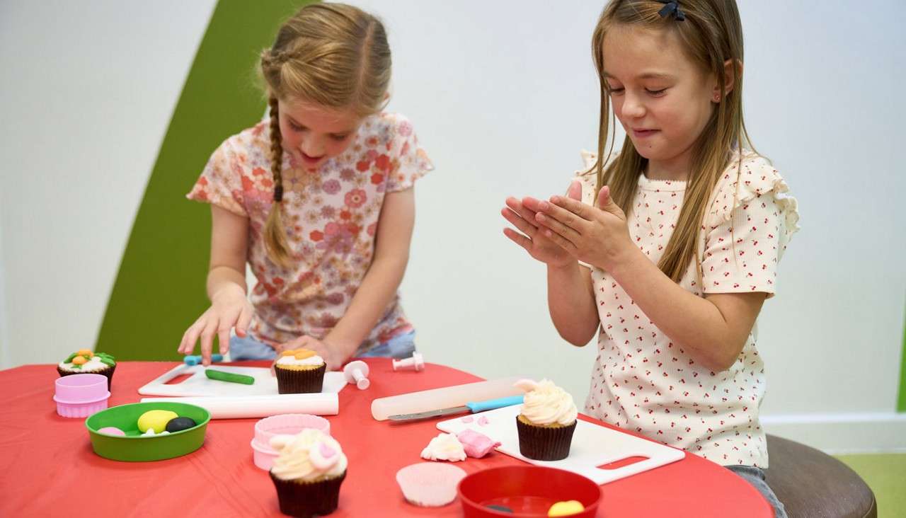 Two children decorate cupcakes, shaping fondant with their hands and small tools. They sit at a red-topped table with cutting boards, bowls, and icing, in a bright indoor classroom.