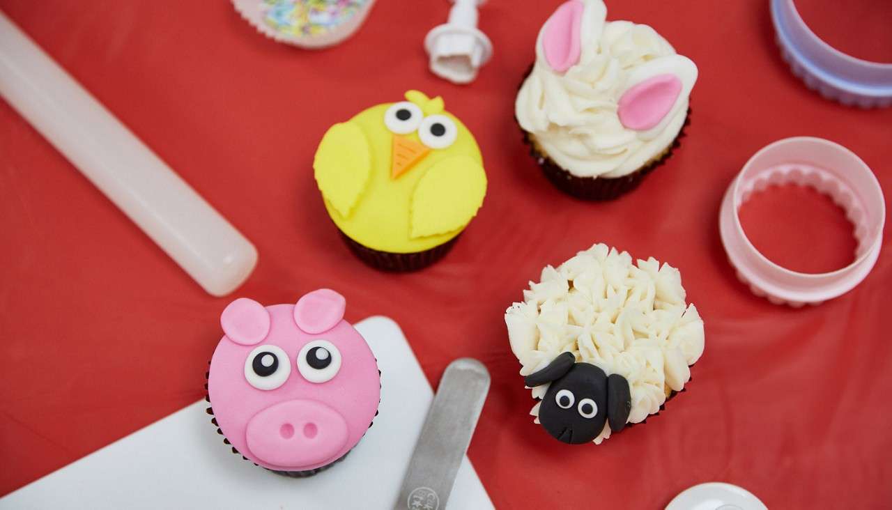 Four animal-themed cupcakes sit arranged on a red tabletop, surrounded by baking tools. A pig, chick, bunny, and sheep decorations rest idle amid cutters, spatula, and sprinkles.