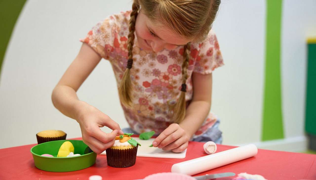 Child with braided hair decorates a cupcake, placing fondant leaves and orange shapes on top, at a red-topped table with bowls, rolling pin, and knife in a brightly colored room.