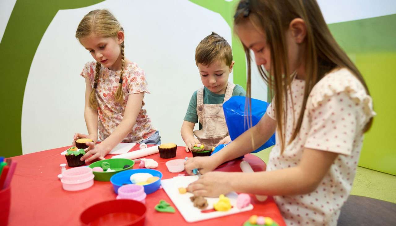 Three children mold colorful playdough, shaping decorations and pretend cupcakes, using cutters and rolling pins, at a red table in a bright classroom with green-and-white walls and scattered bowls.