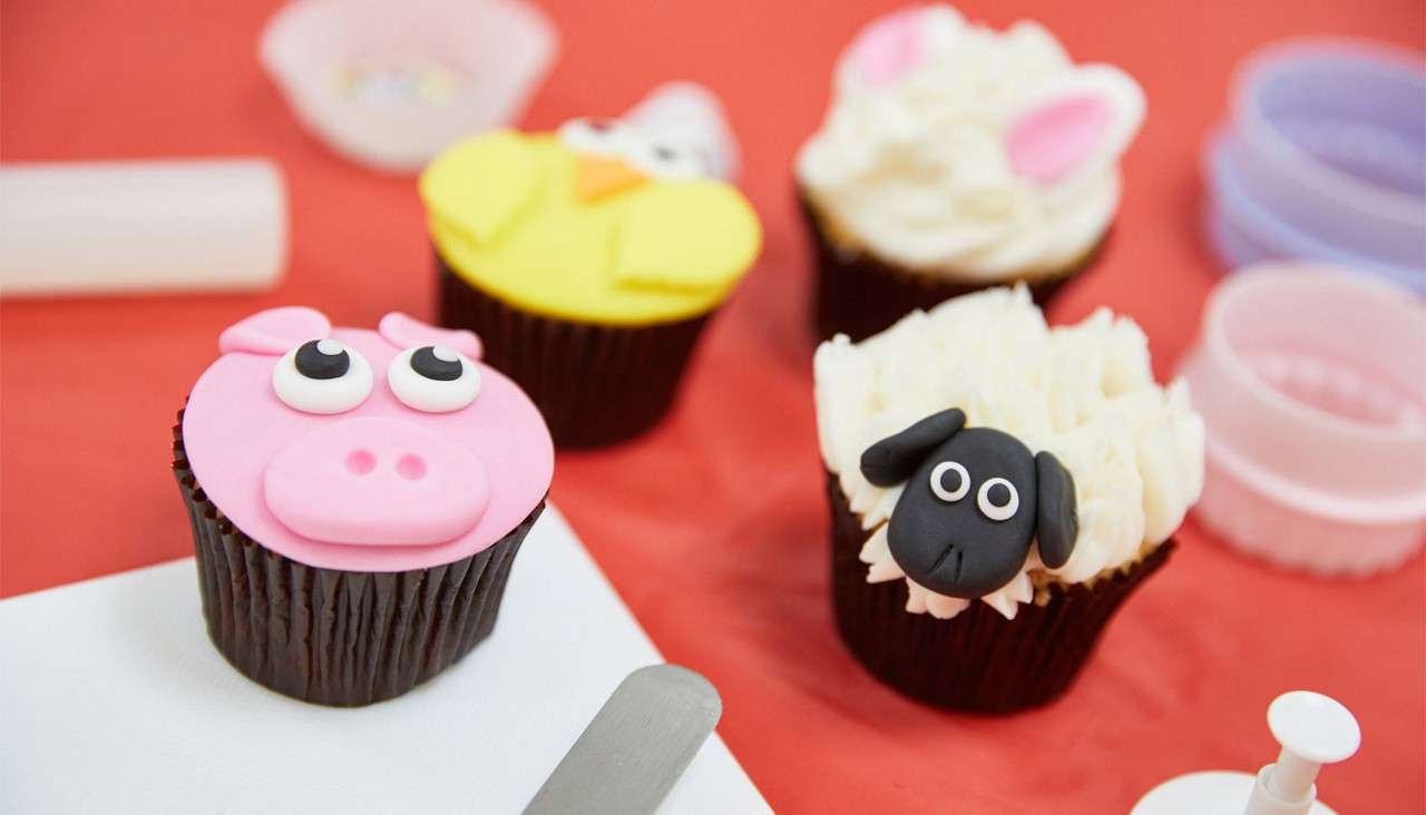 Cupcakes display animal faces, decorated with fondant and frosting; a pig, chick, bunny, and sheep sit on a red tabletop surrounded by cutters and a small spatula during baking prep.