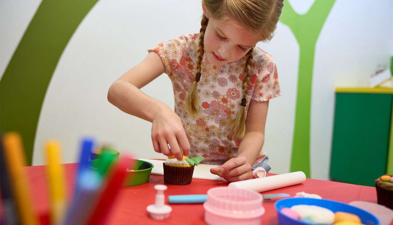 Child decorates a cupcake, placing green fondant leaves on top, at a craft table with baking tools, bowls, and colored pencils in a bright classroom with playful green wall shapes.