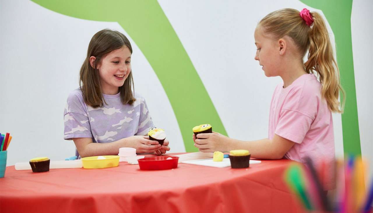 Two children decorate cupcakes, holding frosted treats with yellow icing and candy eyes, while smiling and chatting at a red-covered table in a bright craft room with bowls and tools.
