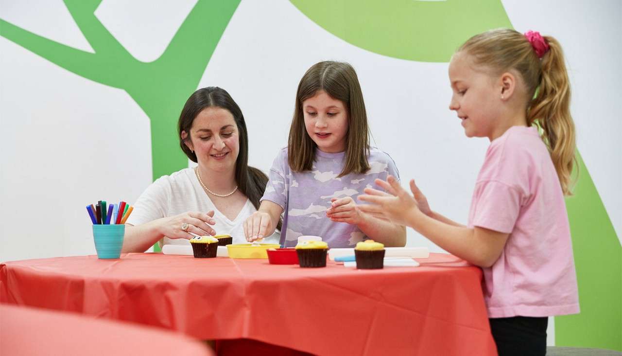Two children and an adult decorate cupcakes, shaping icing with hands at a red table. Cup of markers and trays sit nearby, in a bright classroom with green wall graphics.