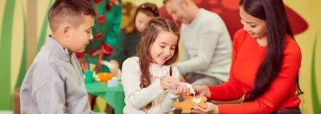 Girl pipes icing onto a gingerbread cookie while a woman holds it; a boy watches nearby, in a colorful classroom with other people crafting at tables.