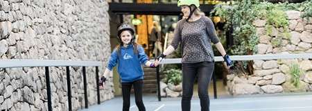 Two helmeted people, an adult and a child, skate hand-in-hand, moving along a smooth blue path bordered by stone walls and railings, approaching a building entrance surrounded by plants.
