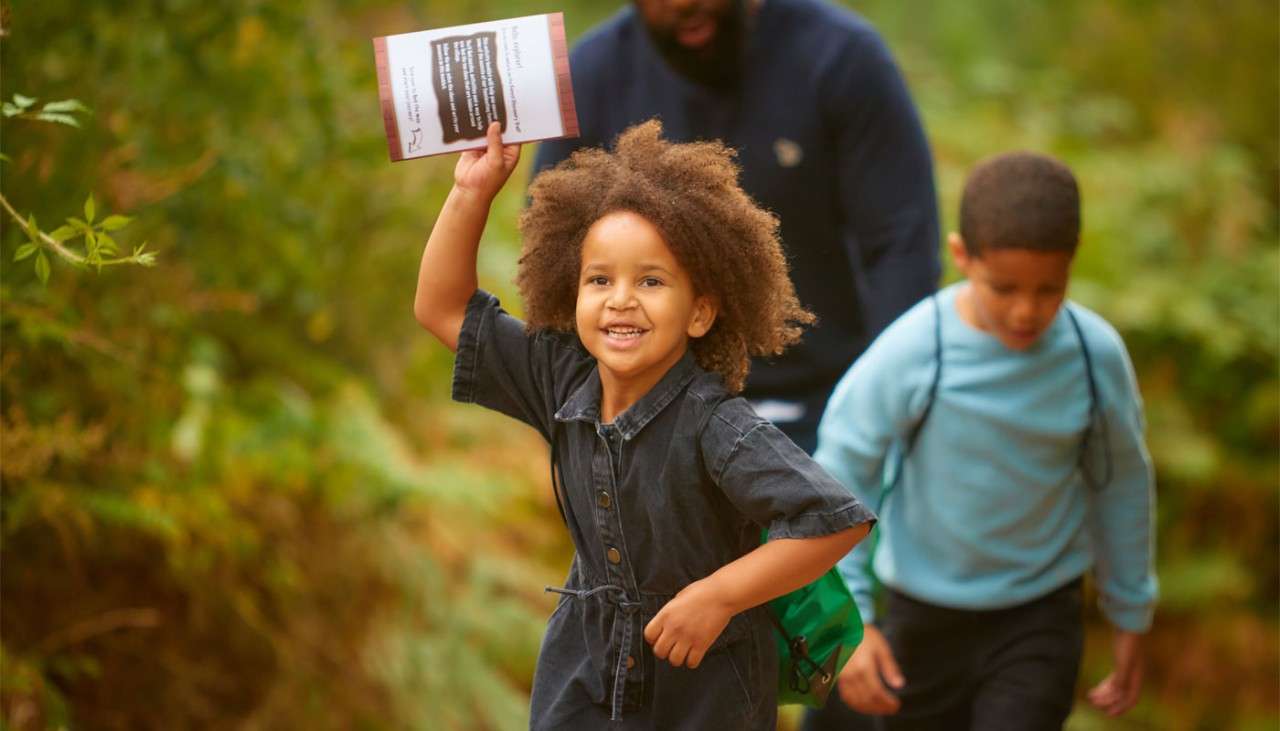 Young girl waving a leaflet whilst running along a woodland path.
