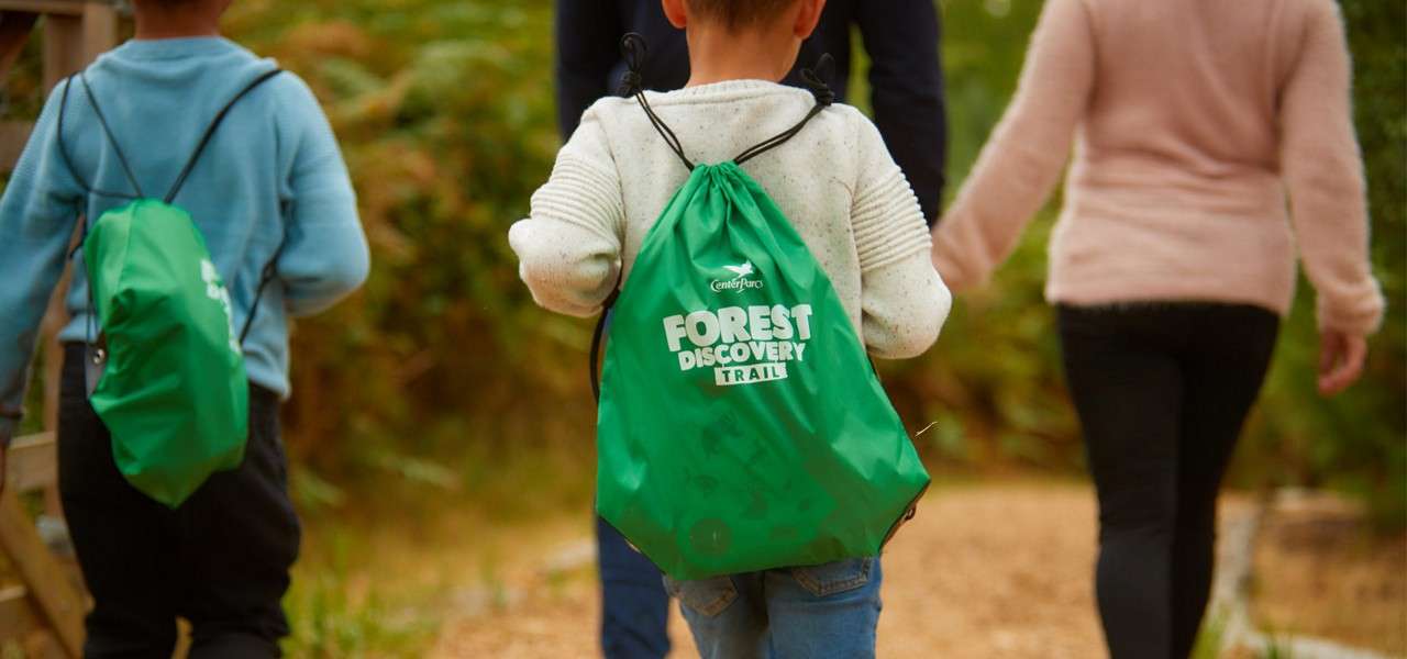 Children wearing green drawstring backpacks walk along a wooded path with adults. Text on bags: “Center Parcs FOREST DISCOVERY TRAIL”.