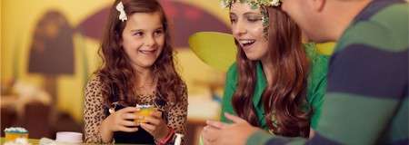 Child and costumed adult decorate a small craft while smiling; a man chats beside them, in a warmly lit party setting with colorful decorations and a soft-focus background.
