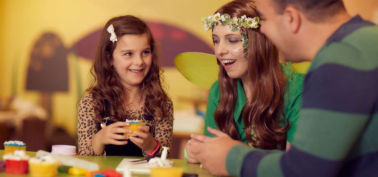 Girl and two adults decorate cupcakes at a craft table, chatting and smiling. One adult wears green fairy wings and a floral crown. Colorful supplies scatter in a party room.