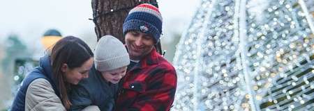 Family trio hugs closely, smiling; adults in winter coats hold a bundled child. Outdoors near a tree, beside glowing holiday lights, warm hats and soft daylight create a festive scene.