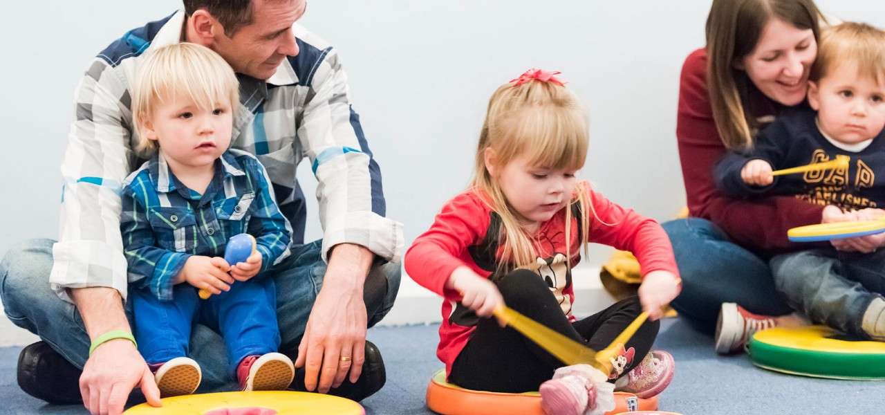 Children play percussion pads with drumsticks while sitting on floor cushions; adults sit beside them supervising in a bright classroom. Visible sweatshirt text: GAP.