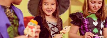 Children in Halloween costumes eat decorated treats on sticks, smiling and chatting, indoors at a party with warm lighting; one wears a witch hat, others wear colorful outfits.