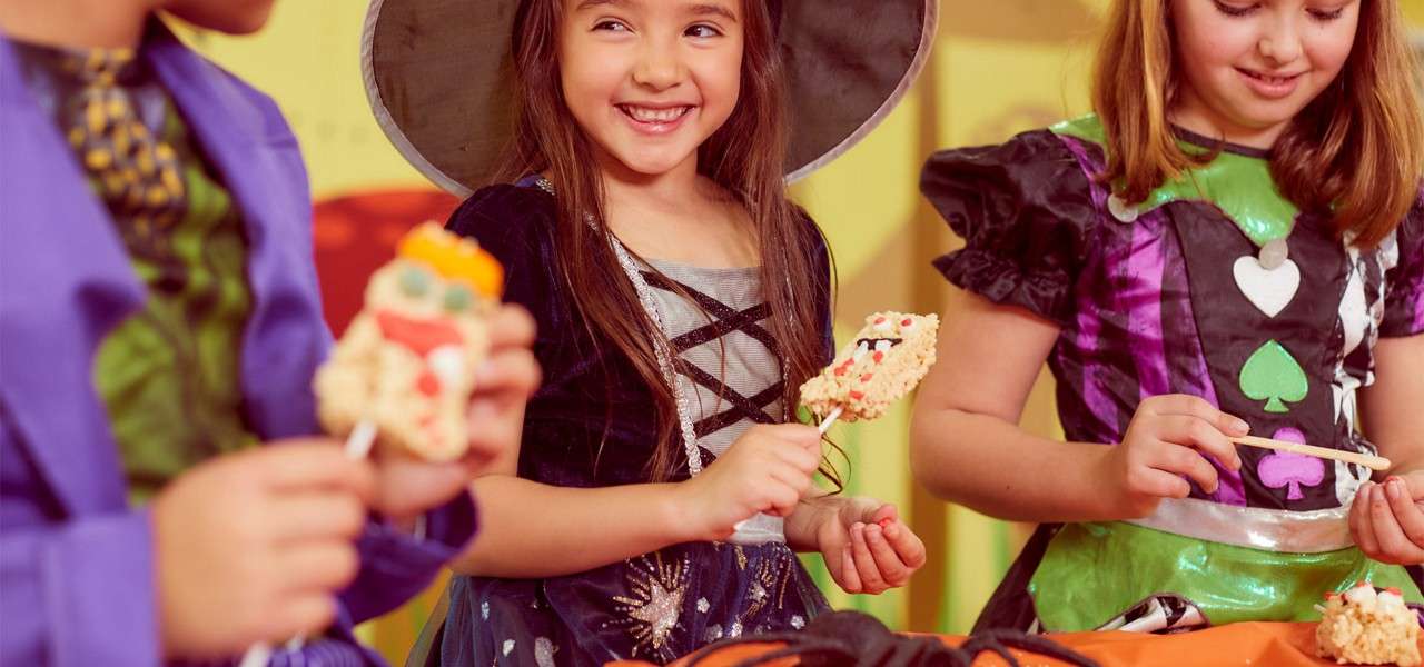 Children decorate rice crispy treat pops, smiling and holding sticks, at a festive party table with orange cloth and pumpkin decorations in a bright indoor room.