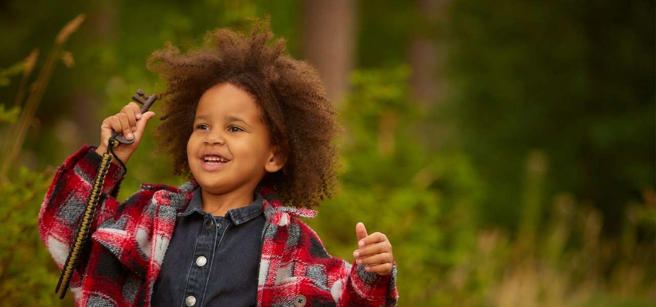 Child smiles and raises a hand while holding a rope handle, wearing a red plaid jacket; outdoors amid green foliage and soft-focus trees on a bright day.