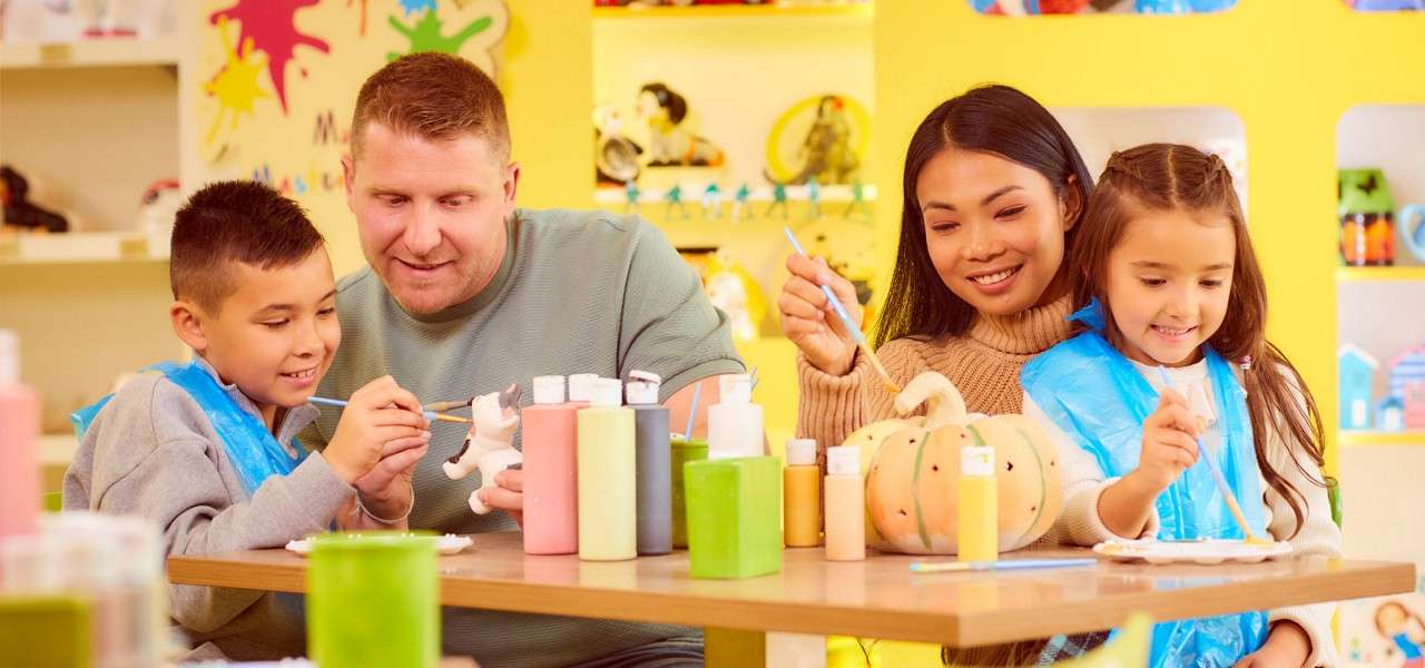 Two adults and two children paint ceramic figurines at a craft table, using brushes and pastel paints; surrounding shelves with toys and yellow walls form a bright studio setting.