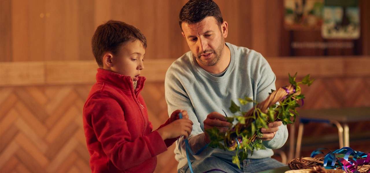 Adult and child weave a leafy wreath, threading blue ribbon through greenery, seated at a table in a warm, wood-paneled room, with craft supplies scattered nearby.