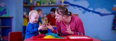 Toddler claps while engaging with colorful sensory noodles; adult mirrors the action, encouraging play. Context: a brightly decorated classroom with shelves of toys and other children in the background.