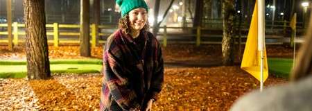 Person in green beanie and plaid jacket smiles, hands clasped, standing on leaf-covered ground at a nighttime mini-golf course, near a yellow flag, trees and fence lit by lamps.