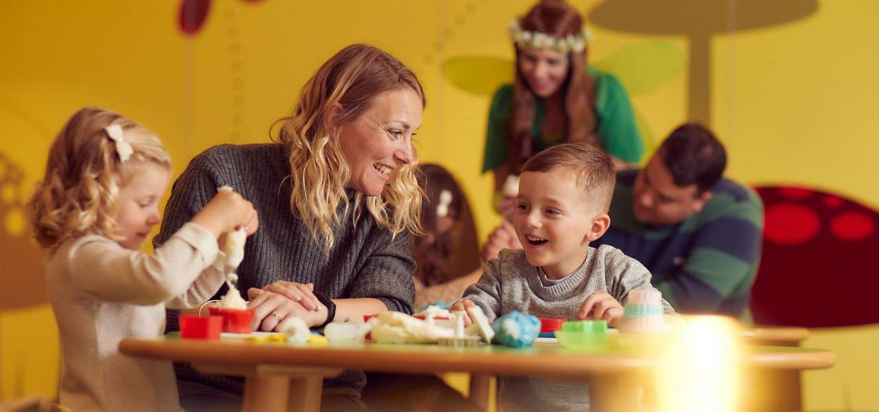 Children craft colorful playdough while a smiling adult guides them at a low table, in a bright, whimsical room with yellow walls, mushroom-like murals, and other adults supervising in background.