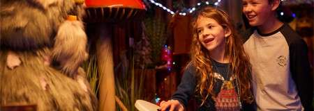 Two children smile and interact with a fuzzy animatronic creature, reaching toward it, in a warmly lit, whimsical room decorated with oversized mushrooms, plants, and colorful string lights.