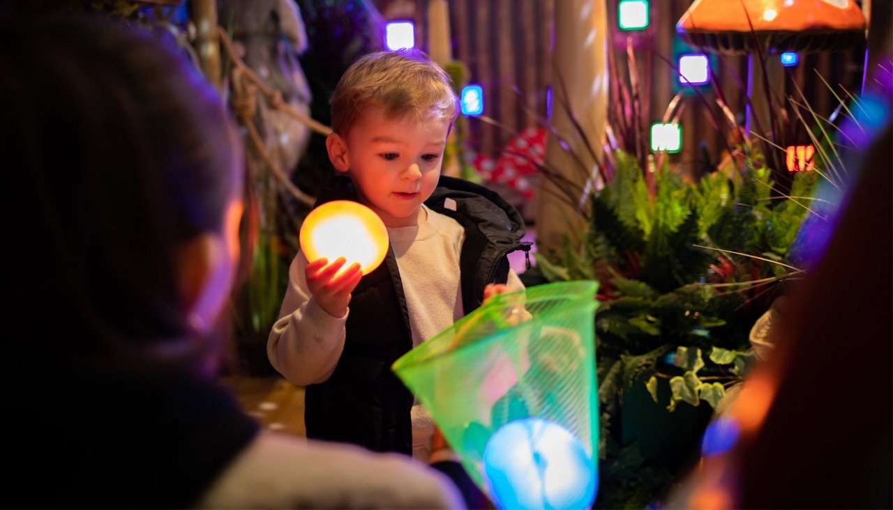 Child holds glowing orb while placing another in a green net, surrounded by colorful lights, square LEDs, plants, and mushroom-like lamps indoors; observer blurred in foreground.