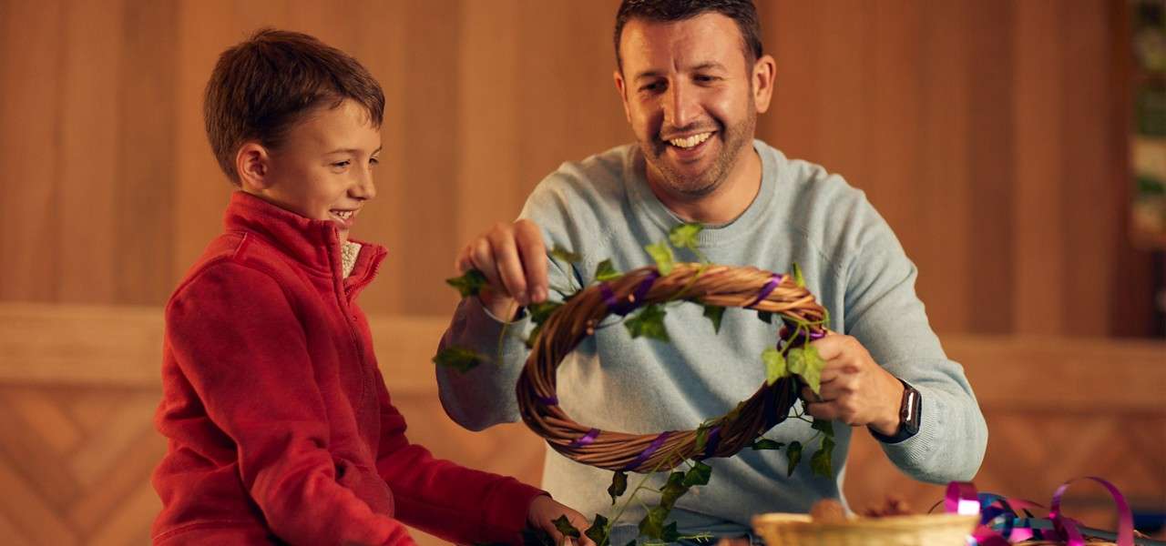 Man and boy weave ivy and purple ribbon into a wicker wreath, smiling and seated at a table; basket and craft materials nearby in a warm, wood-paneled indoor room.