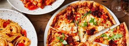 Pizza sits sliced on a plate, melting cheese and herbs visible; nearby, bowls of spaghetti and rigatoni in tomato sauce rest on a wooden table with a glass.