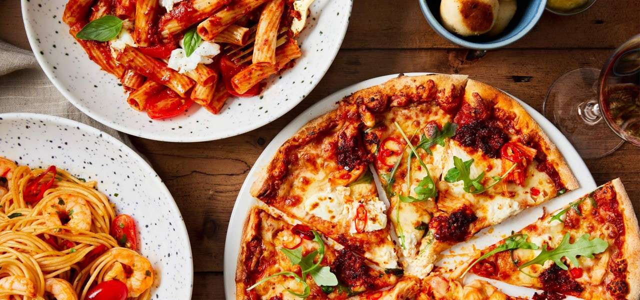 Cheese pizza is sliced and served, with rigatoni and shrimp spaghetti nearby on white plates, with bread rolls and a glass of red wine on a wooden table.