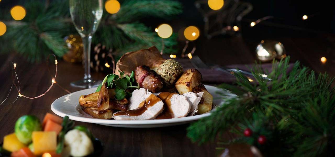 Roast turkey slices with gravy rest on a plate with potatoes, stuffing ball, bacon-wrapped sausage, and greens, on a festive table with pine branches, fairy lights, and a champagne flute.