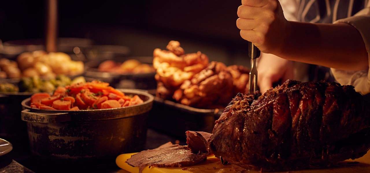 Roast beef is sliced with a carving fork and knife on a cutting board, surrounded by buffet sides—carrots, potatoes, Brussels sprouts, and Yorkshire puddings—under warm, dim lighting.