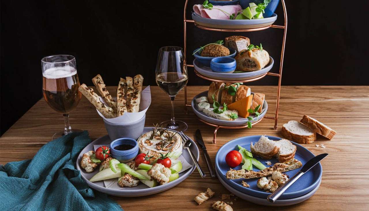 A tiered serving stand presents assorted savory snacks; nearby plates hold bread, vegetables, cheese, and dips. Context: wooden table setting with wine and beer glasses, utensils, and scattered crumbs.