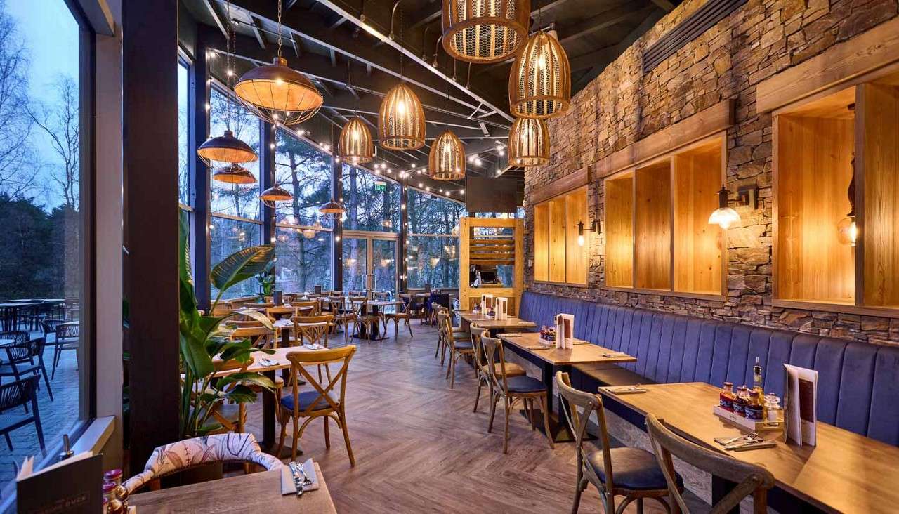 Dining tables sit arranged neatly, awaiting patrons, in a rustic restaurant featuring blue banquette seating, wooden walls, and woven pendant lights, with floor-to-ceiling windows revealing a tree-lined patio at dusk.