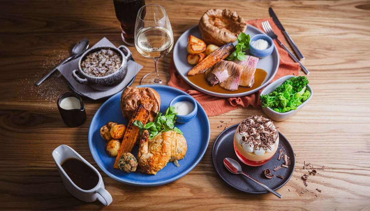 Plated roast dinners sit arranged on a wooden table, featuring meat slices, Yorkshire pudding, roast potatoes, carrots, gravy, sauces, broccoli, a trifle dessert, a powdered dessert, and wine and soda.