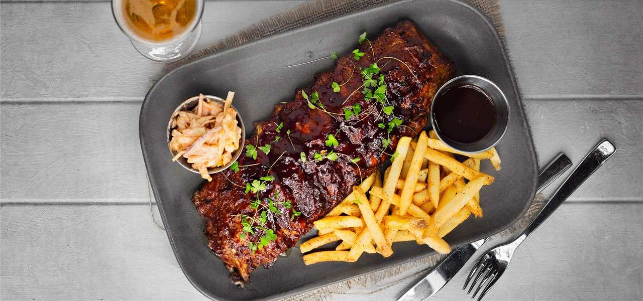 A rack of glazed barbecued ribs rests on a dark platter, garnished with microgreens, accompanied by seasoned fries, coleslaw, and a cup of sauce on a gray tabletop with cutlery.