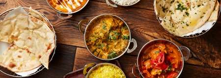 Assorted Indian dishes sit arranged on a wooden table: metal bowls of red and yellow curries, creamy sauce, and saffron rice, alongside baskets of naan and garlic flatbread.