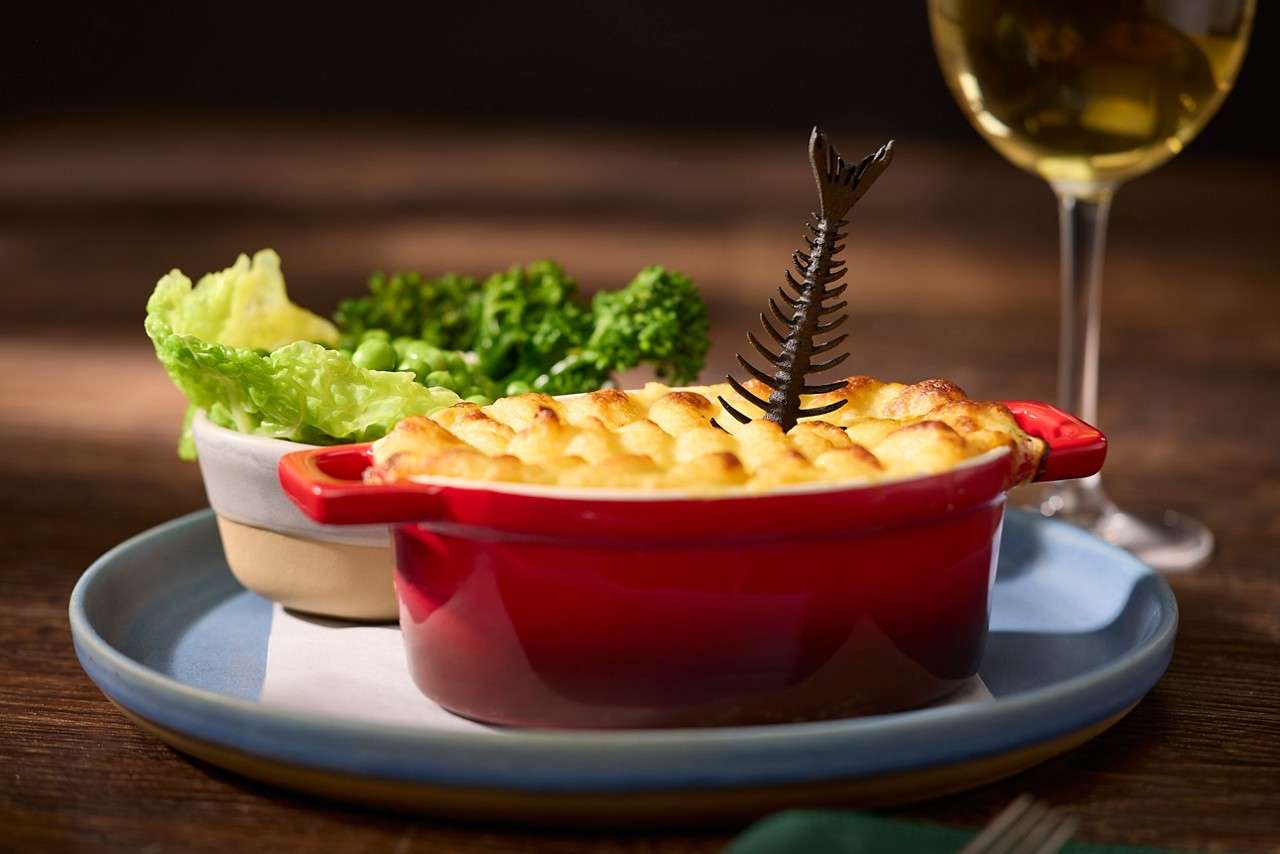 Fish pie in a ceramic dish served with broccoli and a glass of white wine.